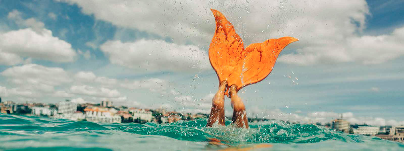 Person playfully wearing an orange mermaid tail fin splashing in the ocean with a cityscape in the background under a bright, cloudy sky.